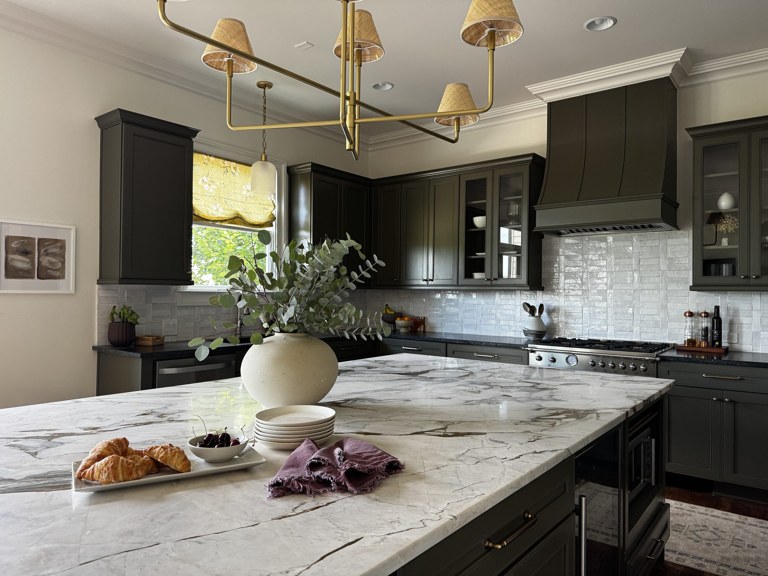 A warm, editorial kitchen designed by Kelly Conrad & Co., with a brass and rattan chandelier above a marble island styled with eucalyptus, croissants, and cherries.