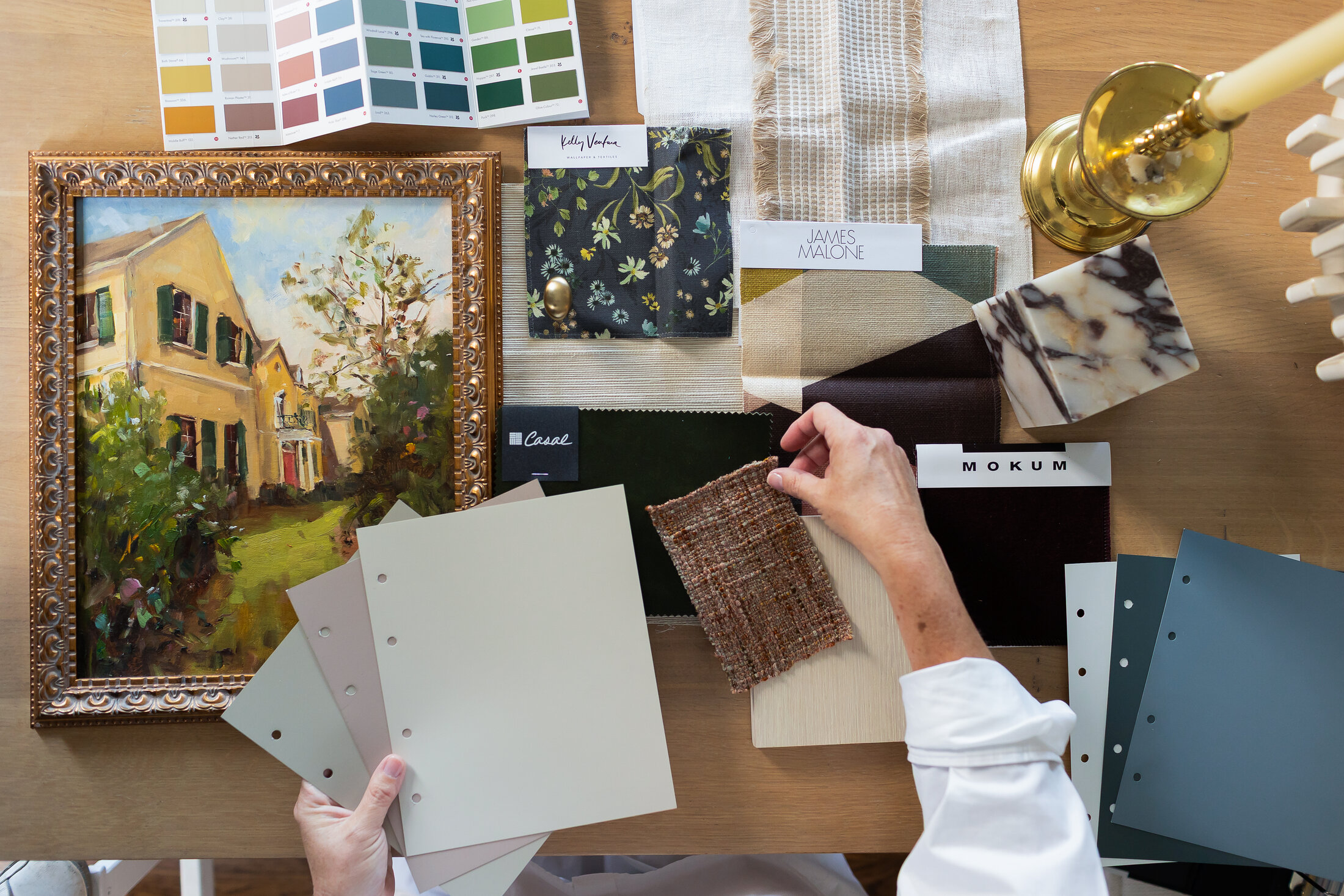 Hands selecting a woven tweed fabric swatch amid paint chips, a framed painting, marble, and a brass candlestick.