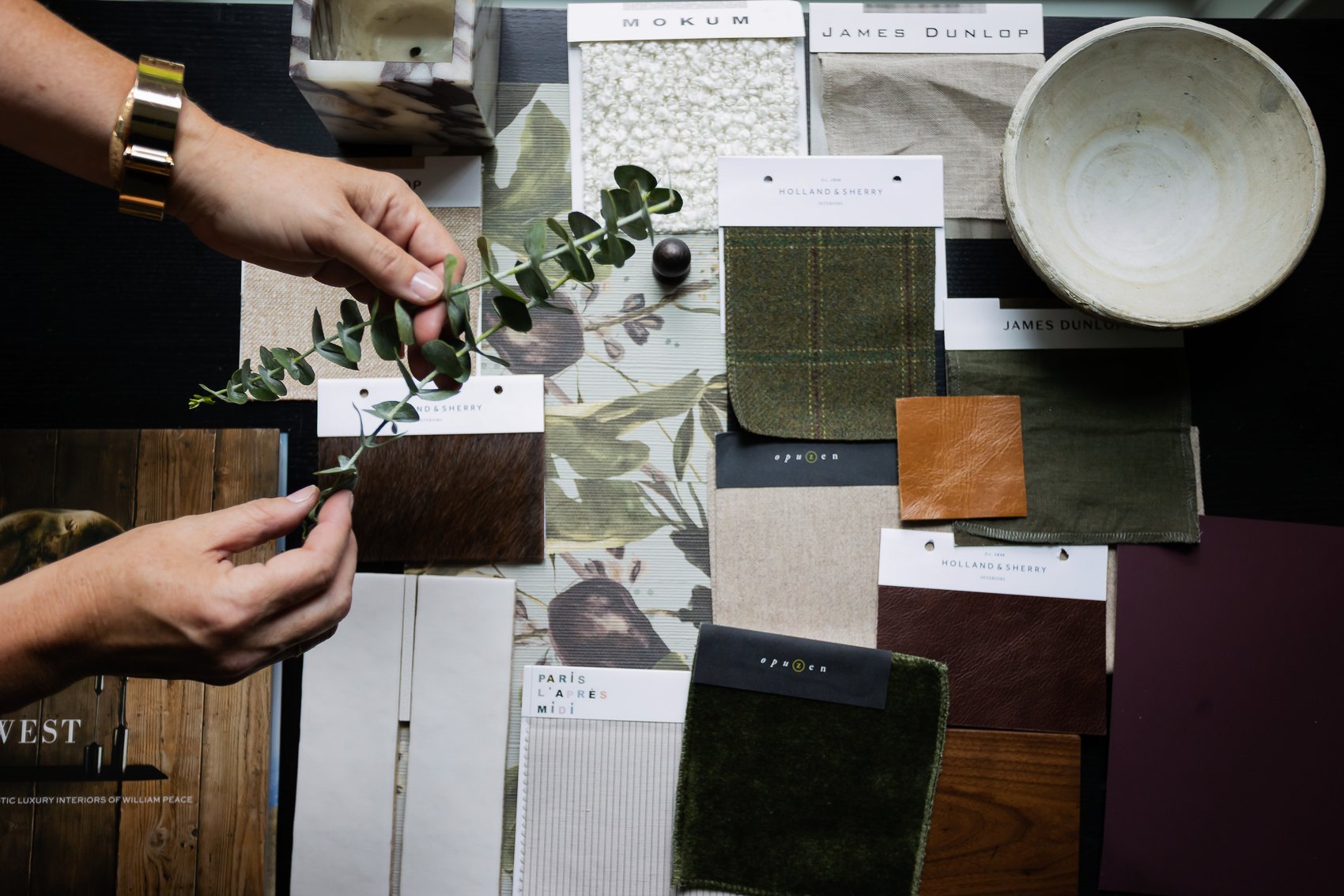 Hands arranging eucalyptus over a design mood board with fabric, leather, and tile swatches.
