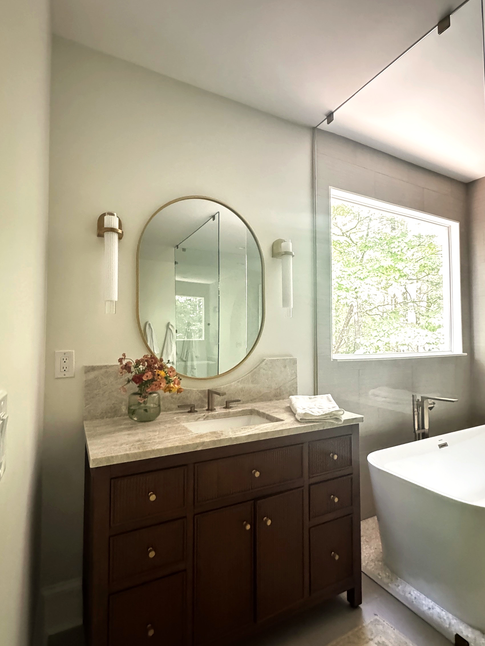 Freestanding soaking tub beneath a window, with vanity in the background.