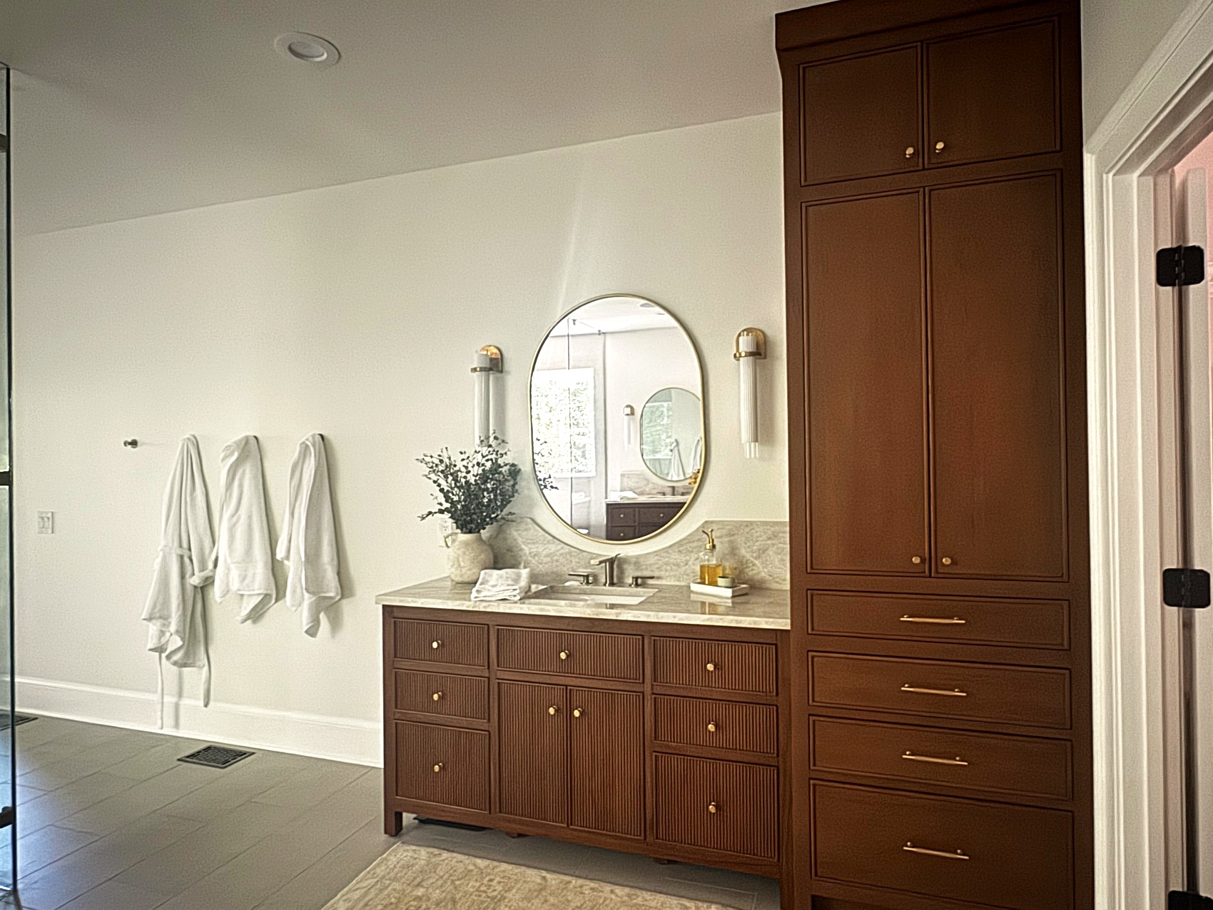 Dark wood fluted vanity with brass oval mirror and tall linen cabinet.