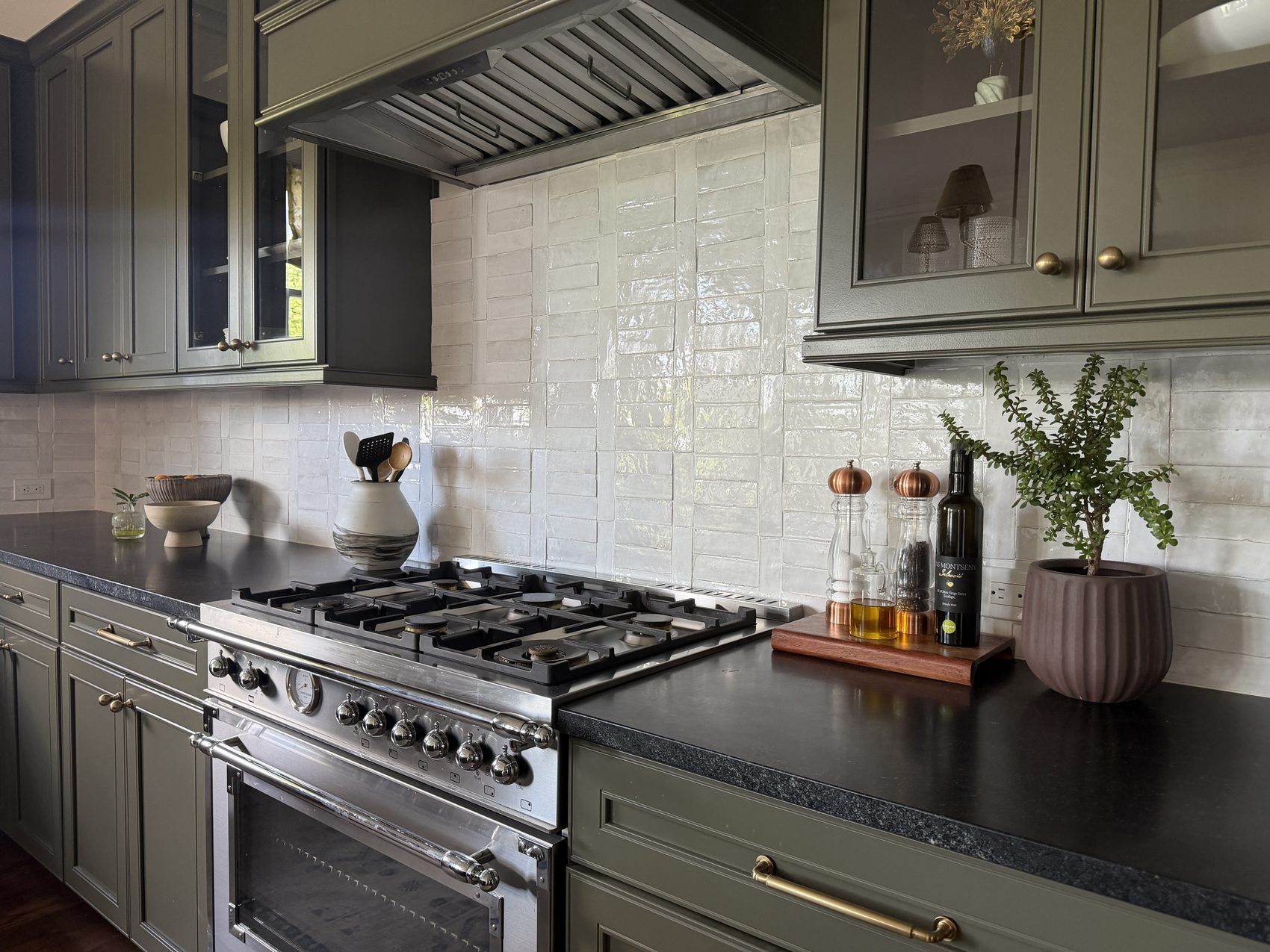 Olive green range hood over gas range with white zellige tile backsplash and styled oil and pepper grinders.