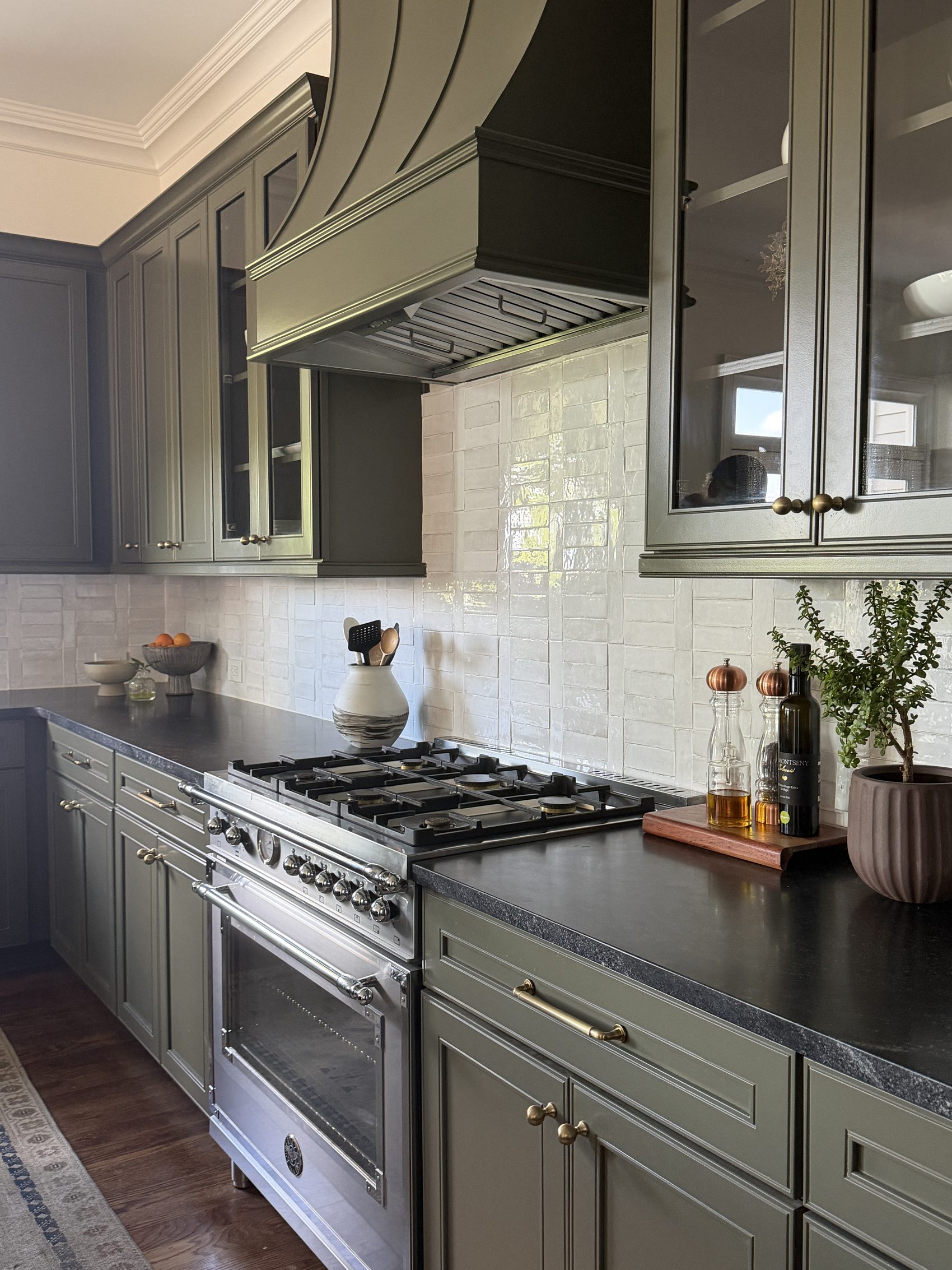 Angled view of olive range hood, gas range, and tile backsplash styled with oil and herbs.