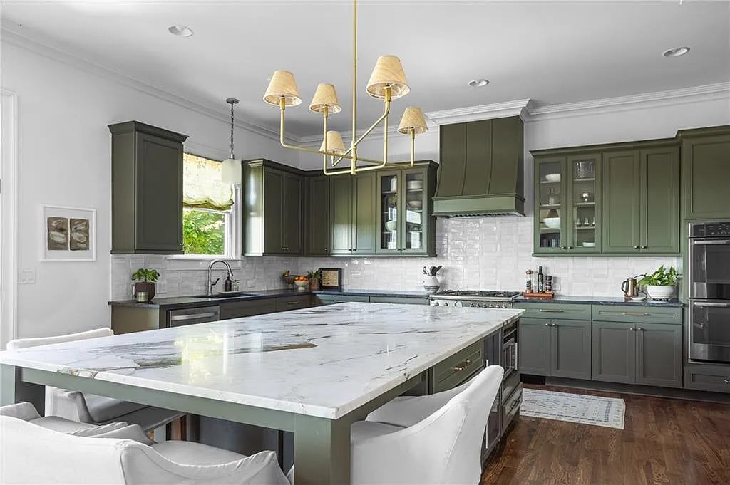 Olive green kitchen viewed from the dining table, with marble island and brass chandelier.
