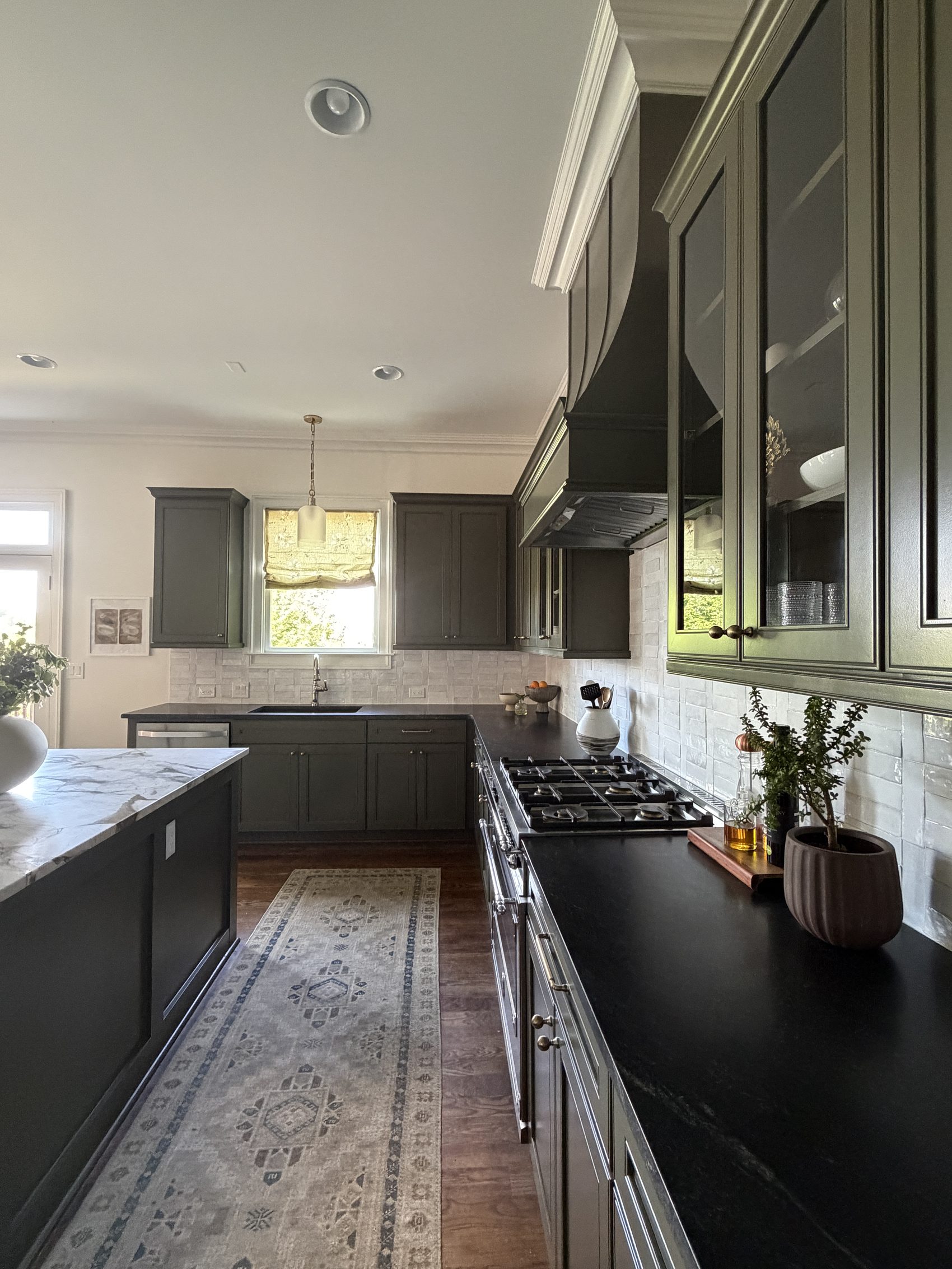 View down the kitchen with a runner rug, marble island, and range beneath a large hood.