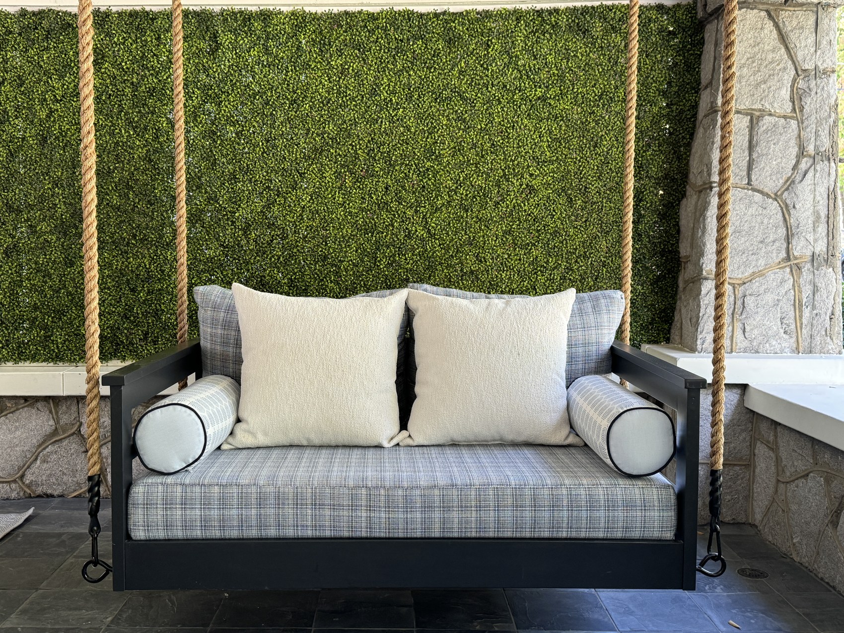 Outdoor porch swing bed styled with plaid and boucle pillows against a boxwood wall.