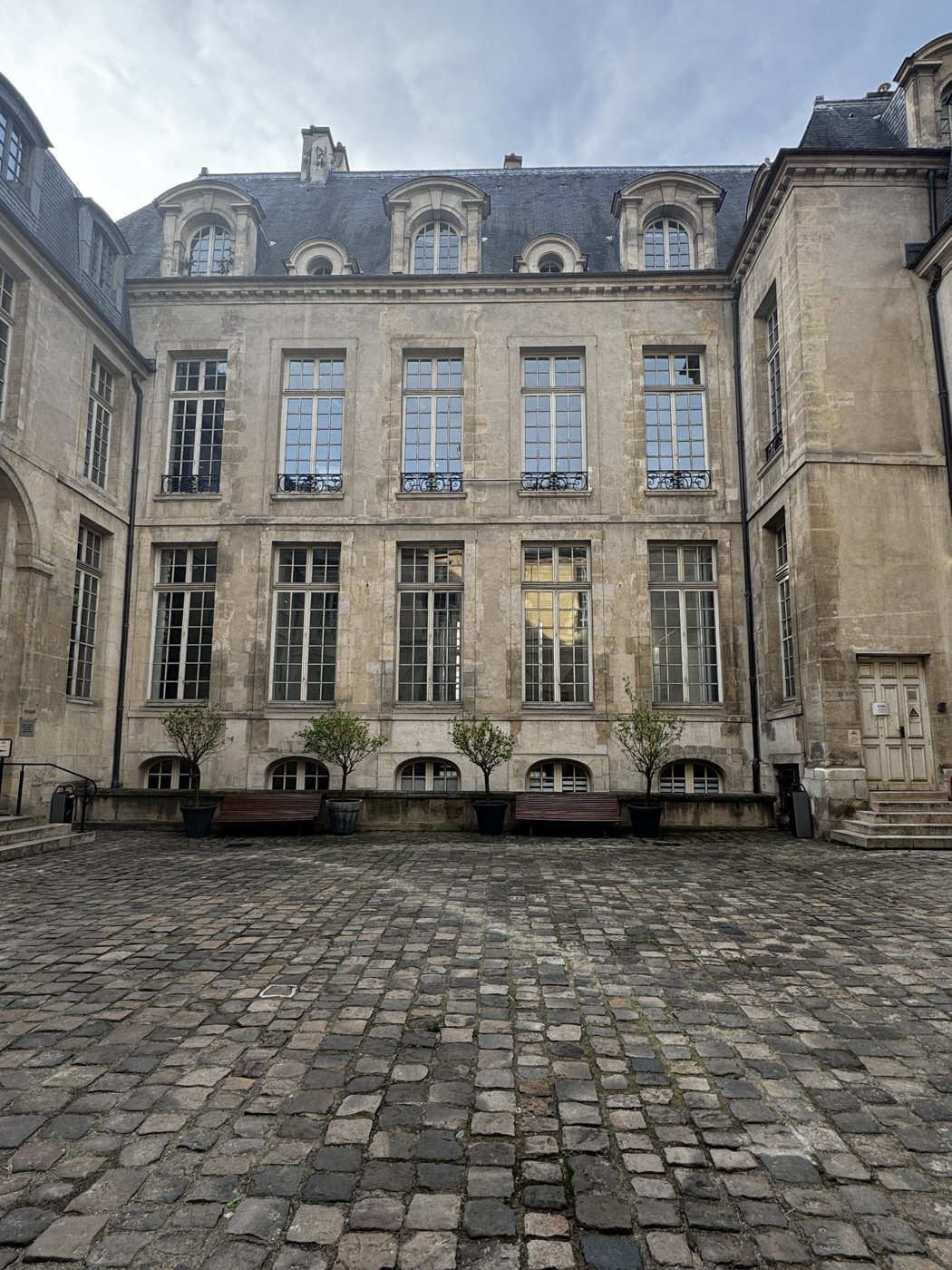 A classic Parisian stone courtyard facade with dormer windows and potted topiaries.