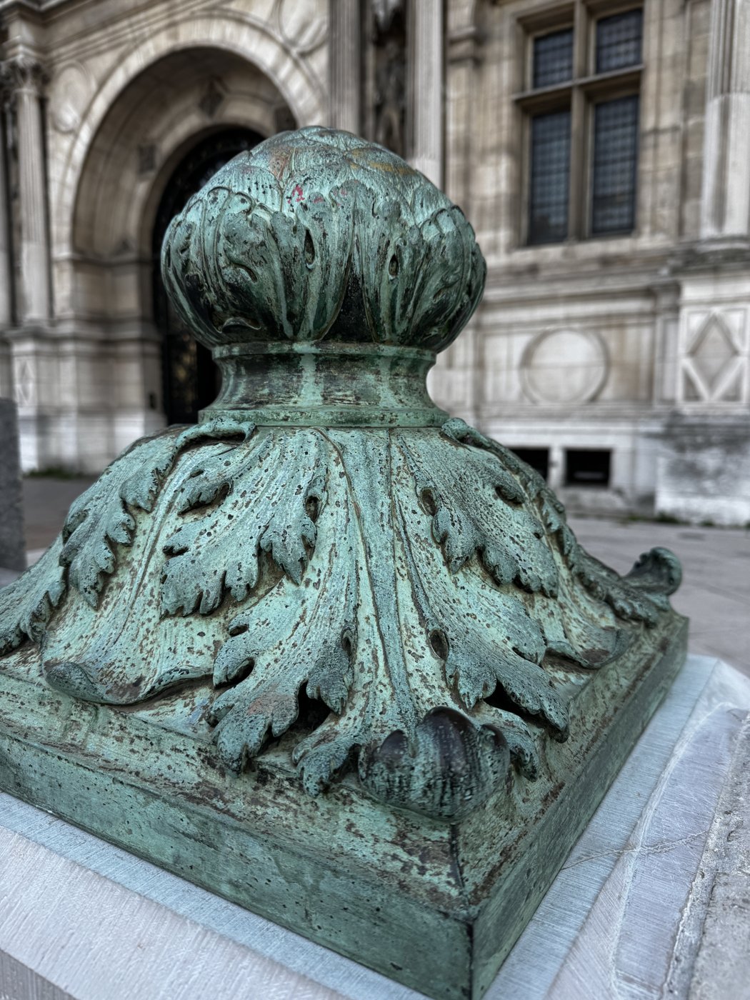 A weathered bronze acanthus-leaf finial in front of a grand stone facade.
